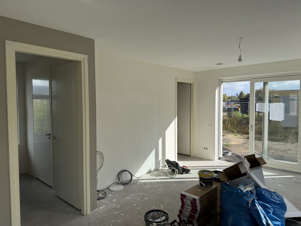 Nieuwbouwwoonkamer tijdens de afwerking, met tuindeuren, verfemmers en dozen op de betonvloer